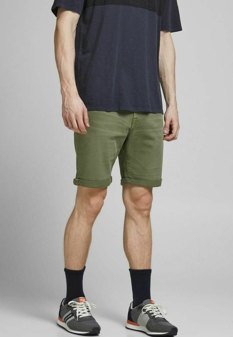 Pantalones cortos de algodón verdes con dobladillos ajustados, combinados con una camisa azul marino y zapatillas grises. Presenta calcetines oscuros y un diseño sencillo.