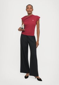 Kirsikkapuna, vartalonmyötäinen yläosa, jossa on hihattomat hihat; leveät, pleated mustat pinstripe-housut; pitää kädessään geometrista hopeista clutch-laukkua; mustat korkokengät.