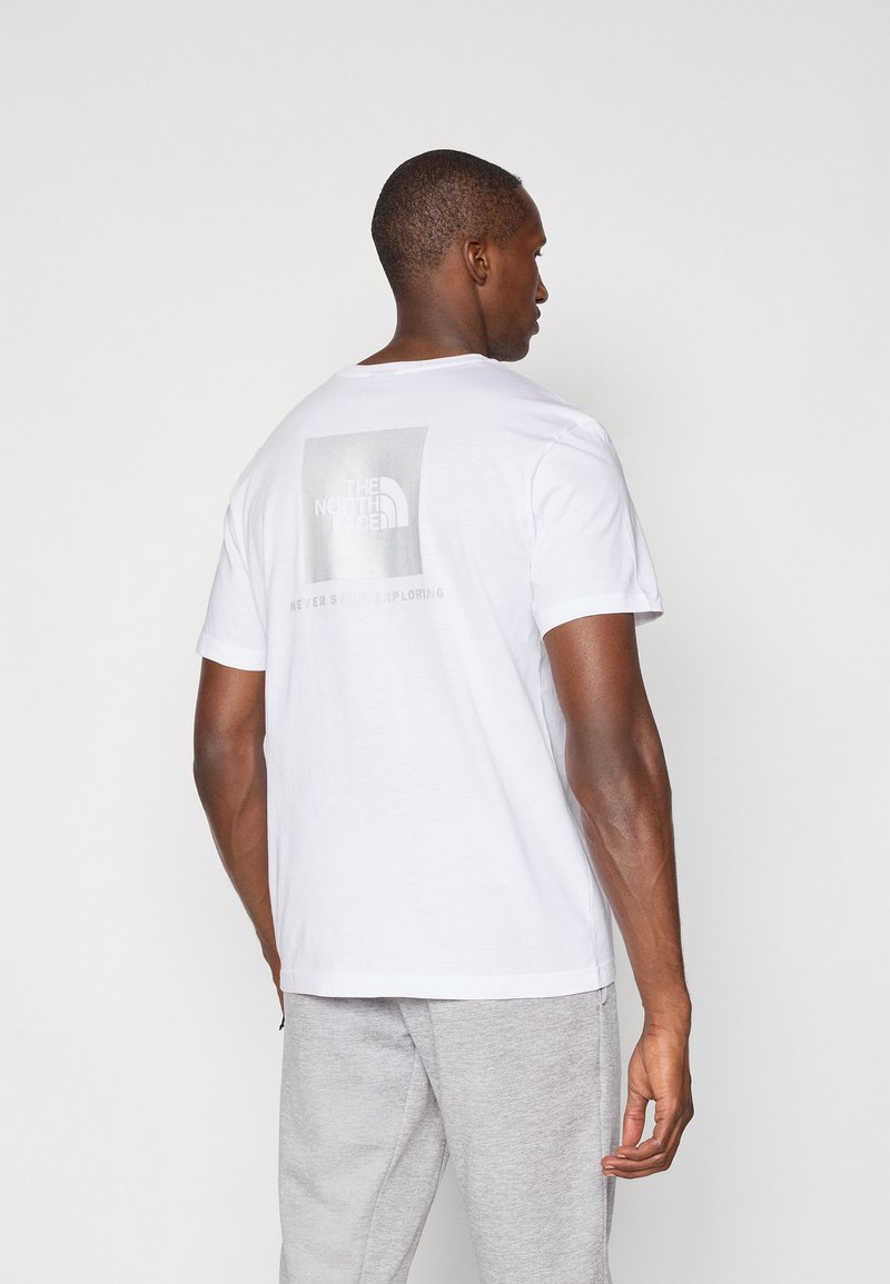 Hvid bomulds T-shirt med korte ærmer, der har et gråt grafisk logo og tekst på ryggen. Sammen med lysegrå sweatpants.