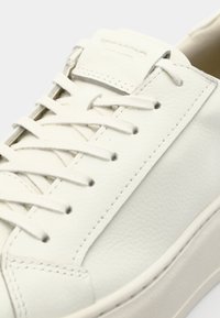 White leather sneaker with textured upper, flat lace closure, and minimalistic design. Features a low silhouette and a rubber sole.