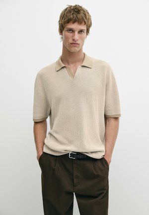Junger Mann mit hellbraunen Haaren trägt beige kurzärmeliges Strick-Poloshirt und dunkelbraune Hose, steht mit den Händen in den Taschen vor weißem Hintergrund.
