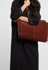 Sac fourre-tout en daim marron avec un design lisse, fermeture éclair et poignées doubles, tenu contre une tenue noire. Silhouette simple et fonctionnelle.