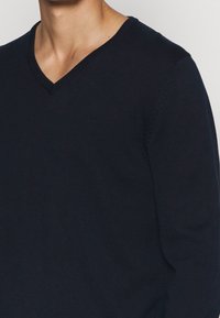 Modellen bär en mörk marinblå V-ringad tröja. Tyget ser jämnt och åtsittande ut, vilket betonar den avslappnade stilen av plagget.