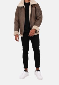 Hnědá šerlinková bunda s krémovým límcem a manžetami, se zipem, nošená přes černou košili, v kombinaci s černými džíny a bílými teniskami.