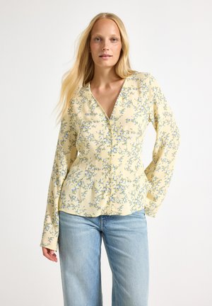 Kvinde med langt blondt hår iført en lys gul blomsterbluse og blå jeans, står mod en almindelig hvid baggrund.
