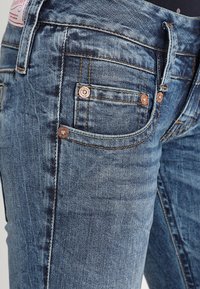 Blå denimjeans med en blekt textur, fem fickor, kopparfärgad hårdvara och synliga sömmar nära midjan.