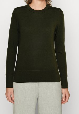 Femme portant un pull ajusté à manches longues vert foncé avec poignets et ourlet côtelés, associé à un pantalon gris clair, sur un fond uni.