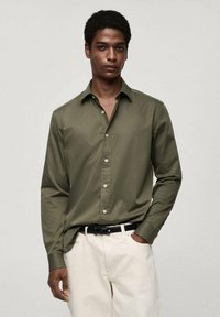 Olijfgroene blouse met lange mouwen, effen stof en een gestructureerde kraag, gedragen met lichte broeken en een zwarte riem.