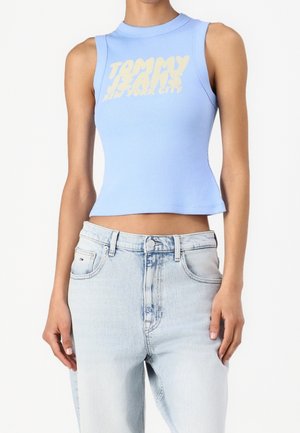 Kobieta nosząca bezrękawnik w jasno-niebieskim kolorze z napisem "Tommy Jeans New York City" oraz jasno-niebieskie dżinsy z wysokim stanem.
