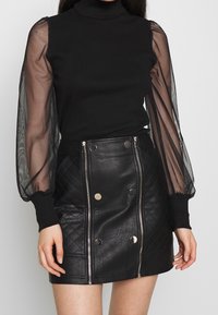 Svart polokrage-topp med genomskinliga puffärmar, i kombination med en svart läder minikjol med quiltad textur och metalliska detaljer.