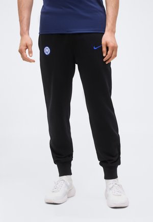 Άνδρας που φοράει μαύρη φόρμα Nike με το λογότυπο της Ίντερ Μιλάνο και μπλε σφιγκόσημο, σε συνδυασμό με λευκά αθλητικά παπούτσια και λευκές κάλτσες.