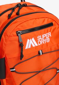 Pomarańczowy plecak z nylonu z podwójnymi zamkami błyskawicznymi, elastycznymi detalami ze sznurka i białym haftowanym logo na przodzie.