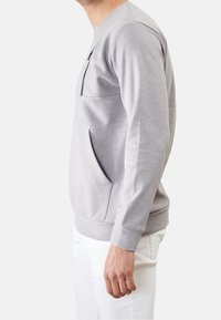 Uomo che indossa un pullover grigio chiaro a maniche lunghe con tasche frontali e pantaloni bianchi, mostrato di lato su uno sfondo bianco.