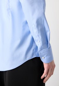 Camisa azul claro con un patrón texturizado, puños con botones y un dobladillo curvado, presentada junto con pantalones negros.