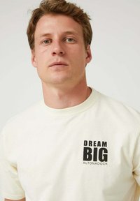 Camiseta de algodón de color crema con mangas cortas y cuello redondo. Gráfico frontal en negro que dice "DREAM BIG ALTONADOCK".