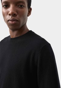Pull en tricot noir avec un col rond, une texture lisse et des manches ajustées. Le tissu semble chaud et doux, adapté pour un usage décontracté.