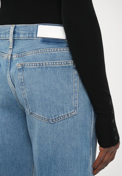 Los jeans de mezclilla azul claro presentan un diseño clásico de cinco bolsillos y una etiqueta blanca en la parte trasera de la cintura. Parte superior negra ajustada con botones en los puños.