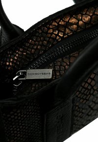 Borsa in pelle nera con finitura testurizzata, dotata di chiusura con zip e un'etichetta metallica impressa con "COWBOYSBAG." Gli accenti includono un leggero luccichio.