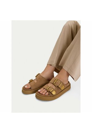 Nohy obuté v béžových sandálech s dvěma velkými zlatými přezkami a silnými podrážkami, doplněné o hnědé kalhoty s širokými nohavicemi.