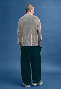 Pruhovaný pletený svetr v béžové a krémové barvě, oversized s rukávy raglán, spárovaný s tmavě zelenými širokými kalhotami a šedými teniskami.