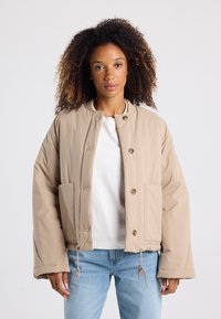 Beige padded jacket with a round collar, front snap buttons, two side pockets, adjustable drawstrings at the hem, and a smooth texture.
