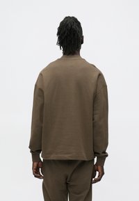 Persona con dreadlocks legati indossa una felpa marrone a maniche lunghe e pantaloni abbinati, in piedi di fronte a uno sfondo bianco uniforme.