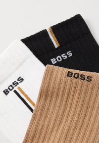 Trois paires de chaussettes : blanches avec une rayure noire et dorée, noires avec une rayure dorée, et beige côtelé. Toutes présentent le logo "BOSS".