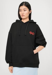 Jovem mulher vestindo um moletom preto com capuz e o logotipo vermelho "Fiorucci", com uma mão no bolso, de pé contra um fundo claro e simples.