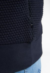 Marineblå striktrøje med et tekstureret mønster, ribbestrikket kant og et lille mærke på siden, parret med lyseblå denim.