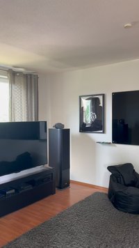 Fekete televízió egy sötét fa média egységen, mellette álló hangfal, puha textúrájú fekete babzsák, világos fa padló.