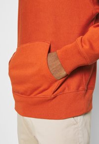 Felpa arancione con polsini e orlo a costine, tasca a marsupio e texture morbida. Il tessuto sembra resistente e confortevole.
