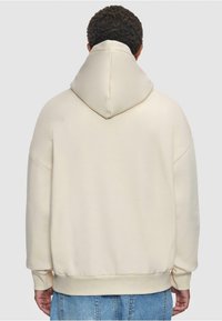 Sweat à capuche crème avec un col montant et une coupe surdimensionnée. Texture douce, poignets et ourlet côtelés, sans logos ni motifs visibles.