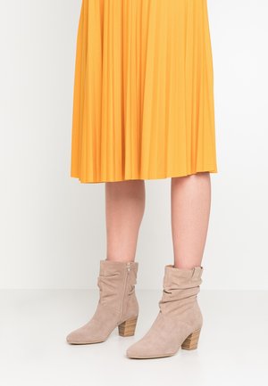 Person iført beige ruskind ankleboots med blokhæle og gul plisseret midinederdel, der står på et hvidt gulv.