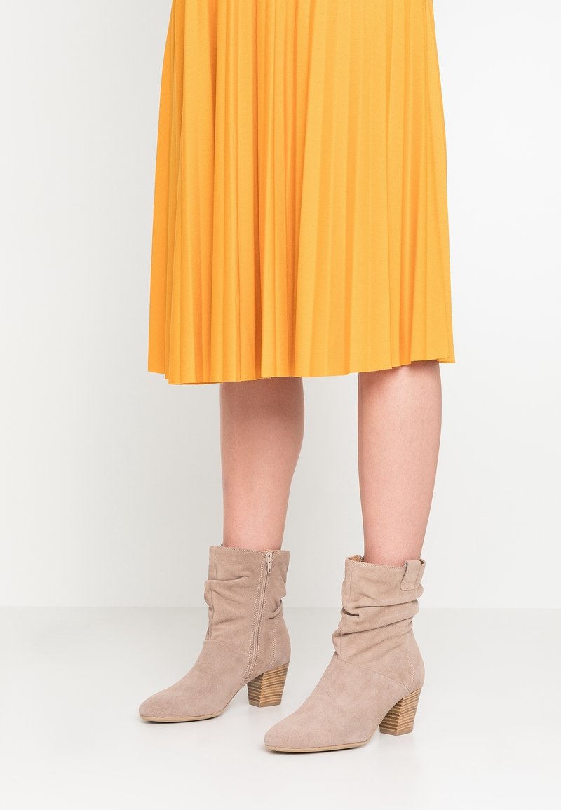 Personne portant des bottines beiges en daim à talons blocs et une jupe midi plissée jaune, debout sur un sol blanc.