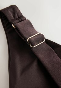 Sangle en tissu marron foncé réglable avec une boucle coulissante en métal argenté sur un fond de vêtement lisse et sombre.