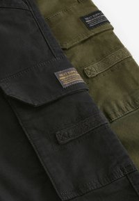 Deux panneaux en tissu : un noir, un vert olive. Les deux comportent des poches, des détails de couture et des étiquettes avec la marque "NEXT SUPPLY".