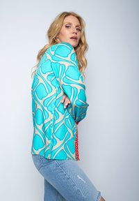 Koszula z długim rękawem w kolorze turkusowym z beżowym abstrakcyjnym wzorem i czerwonymi akcentami. Lekki materiał o gładkiej teksturze oraz luźnym kroju.