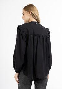 Sort bluse med flæser på skuldrene, samlet detalje på ryggen og lange ærmer. Fremstillet af letvægtsstof med en afslappet pasform.