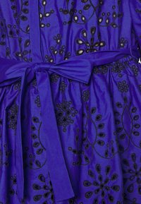 Tissu violet avec broderie florale noire, taille nouée et finition texturée avec des détails découpés dans tout le design.