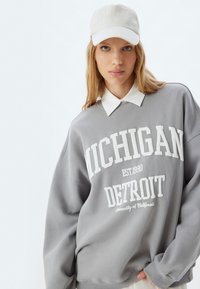 Grauer oversized Sweatshirt mit dem Text "MICHIGAN DETROIT EST 1990" in Weiß. Kombiniert mit einer hellfarbigen Kappe, die den Text "NATURAL" zeigt.
