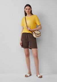 T-shirt en coton jaune avec imprimé palmier, shorts marron à taille élastique, sandales blanches à enfiler et un petit sac bandoulière crème avec des accents marron.