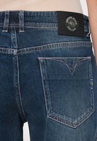 Tmavomodré rifle z denimu s ružovým prešívaním, so štítkom z kože s vytlačeným logom a klasickým dizajnom zadného vrecka.