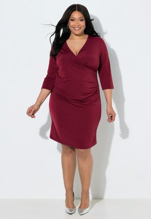 Mujer con cabello largo y oscuro, vestida con un vestido cruzado ajustado burdeos con mangas tres cuartos y tacones altos plateados, de pie contra una pared blanca.