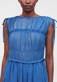 Femme portant une robe bleue transparente sans manches avec des détails à volants et des bretelles nouées aux épaules, montrée du cou à la taille.