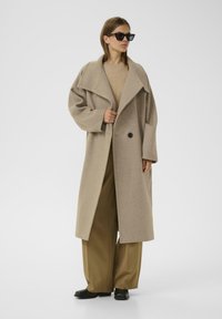 Cappotto in misto lana beige con ampio colletto, due bottoni e vestibilità rilassata. Abbinato a un top beige, pantaloni verde oliva e scarpe nere.