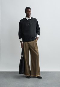 Melns džemperis ar baltu izšuvumu, virsū uz baltas garās piedurknes krekla. Brūnas platajās bikses un melna soma. Gludas tekstūras.