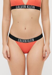Calvin Klein bikini-set i en ljus korallfärgad ribbad tyg, djup V-ringad bh med en svart logoband, samt matchande svart midjeband på trosorna.