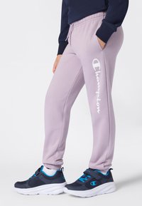 Person klädd i lila Champion sweatpants och marinblå sneakers med blå detaljer, stående med ena handen i fickan.