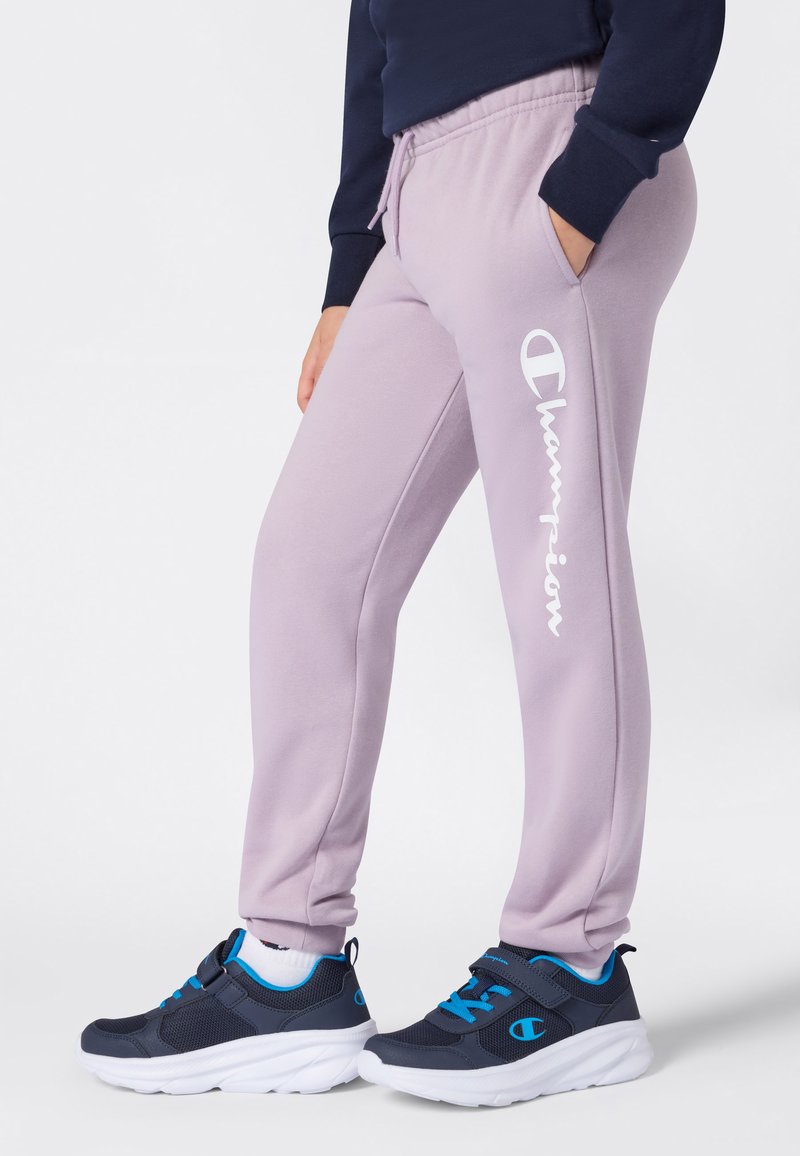 Person klädd i lila Champion sweatpants och marinblå sneakers med blå detaljer, stående med ena handen i fickan.