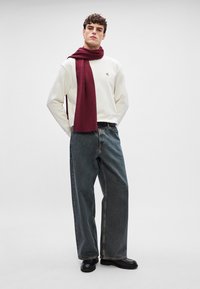 Bílá mikina s malým logem, vínová žebrovaná šála, široké tmavé džíny a černé boty. Jednoduchý design, neformální styl.